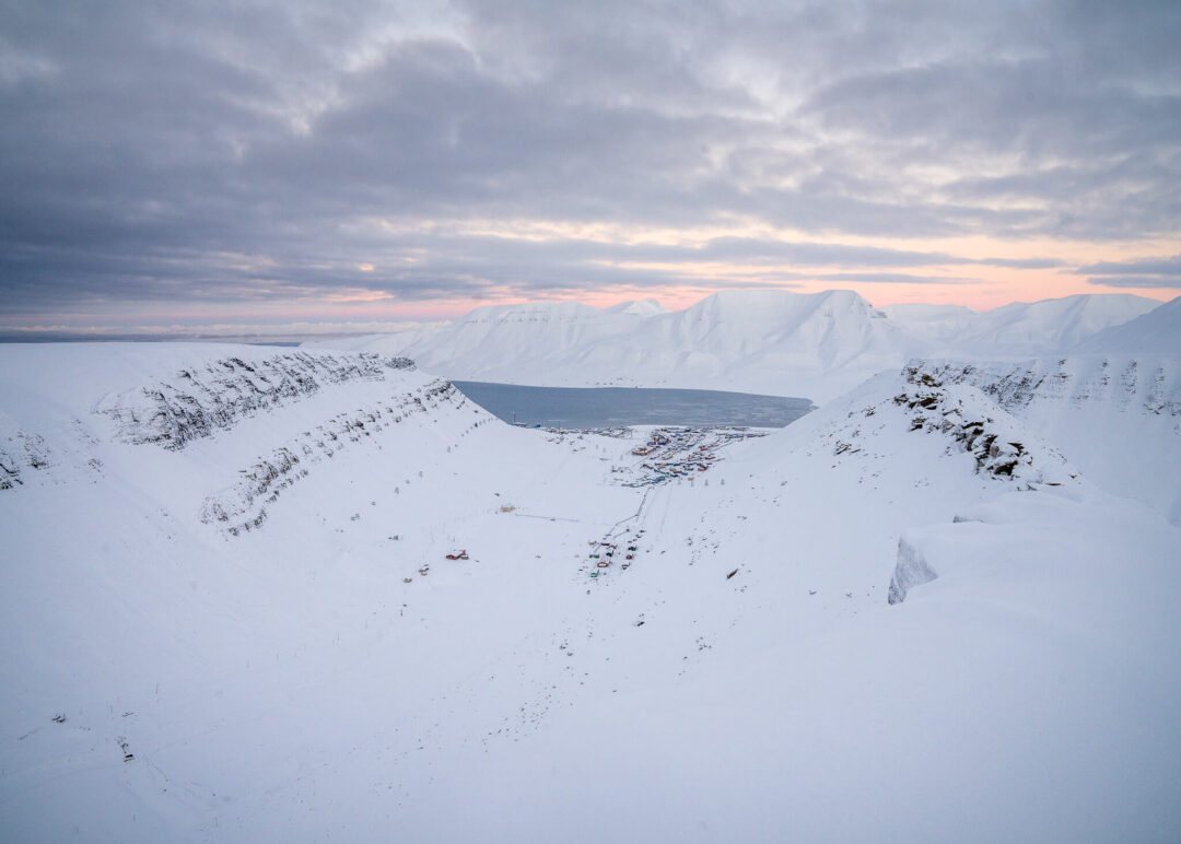 Snowy Svalbard landscape at dusk with frozen fjord and snowmobile tracks across the mountains