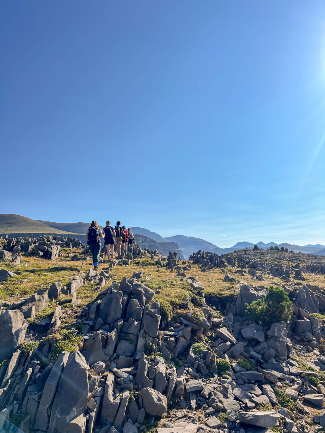 Hikers walking along a rocky mountain trail under a clear blue sky.