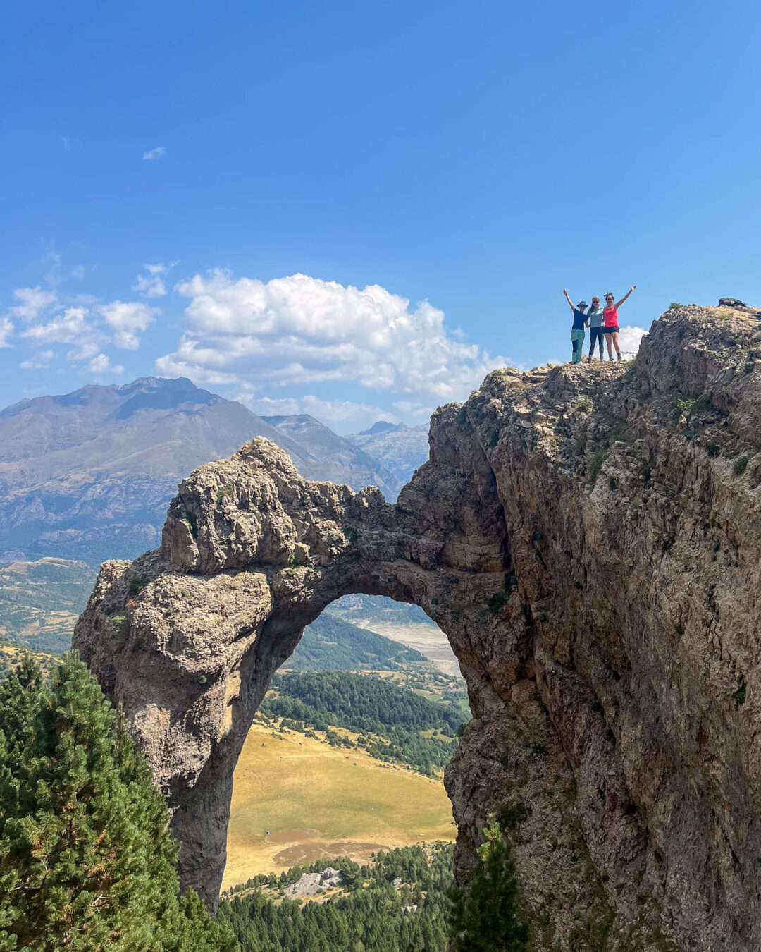 Hikers standing on a rocky cliff by a natural rock arch with sweeping mountain views on a sunny day
