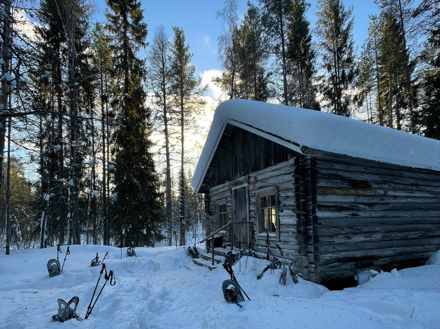 Snow-dusted log cabin in a Finnish forest with snowshoes and cross-country ski gear at dusk
