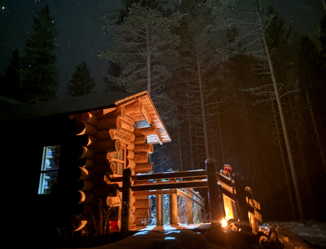 Illuminated wooden log cabin and snowy forest path at night, winter adventure setting