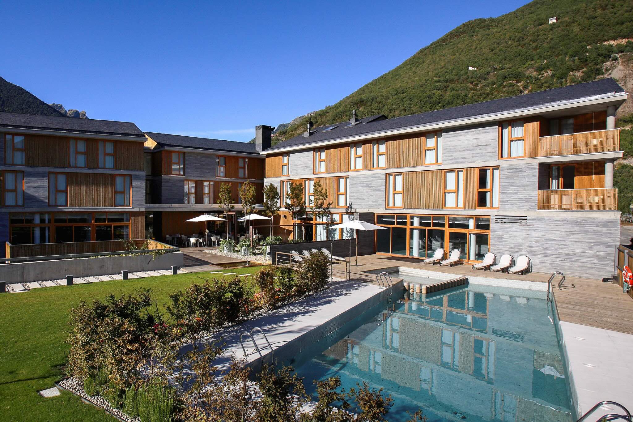 Hotel courtyard with a swimming pool and sun loungers, set against surrounding mountains