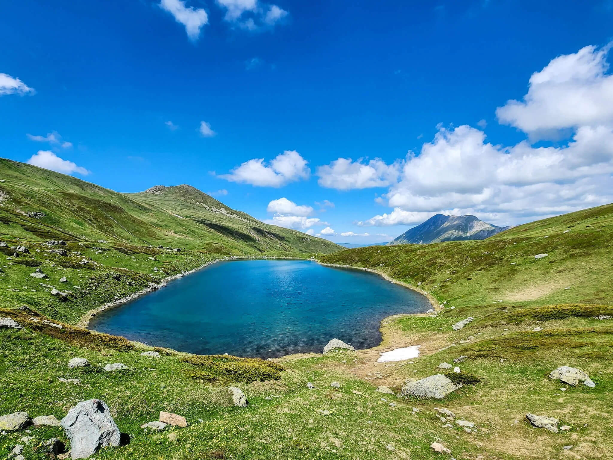 Alpine mountain lake in a green valley with hiking trails under blue sky and clouds