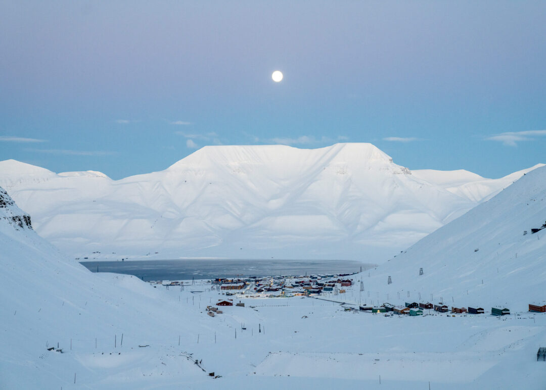 Snow-covered Svalbard harbour settlement with boats and mountains under a clear blue sky