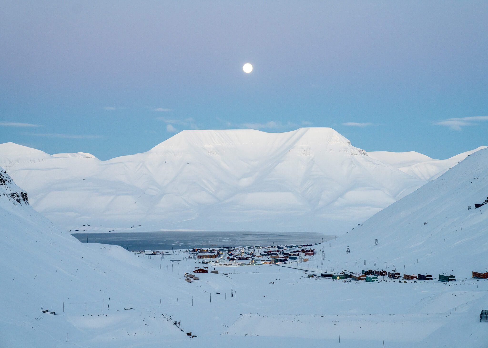 Snow-covered Svalbard harbour settlement with boats and mountains under a clear blue sky
