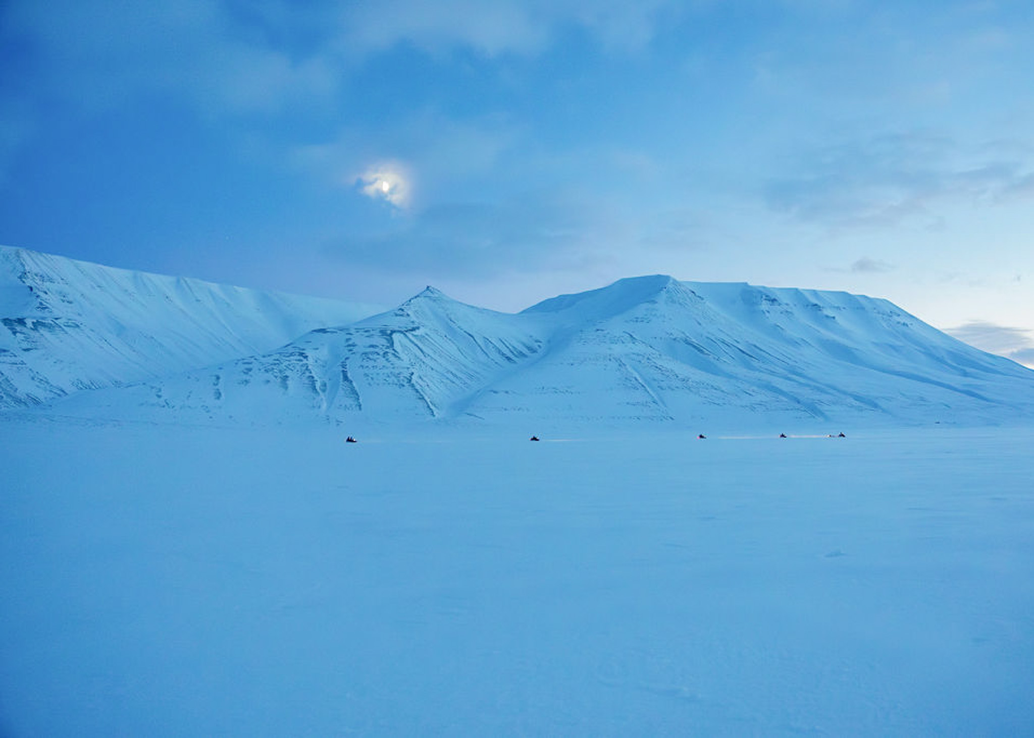 Snow-covered mountains and open Arctic snow landscape with snowmobile tracks across Svalbard under blue sky