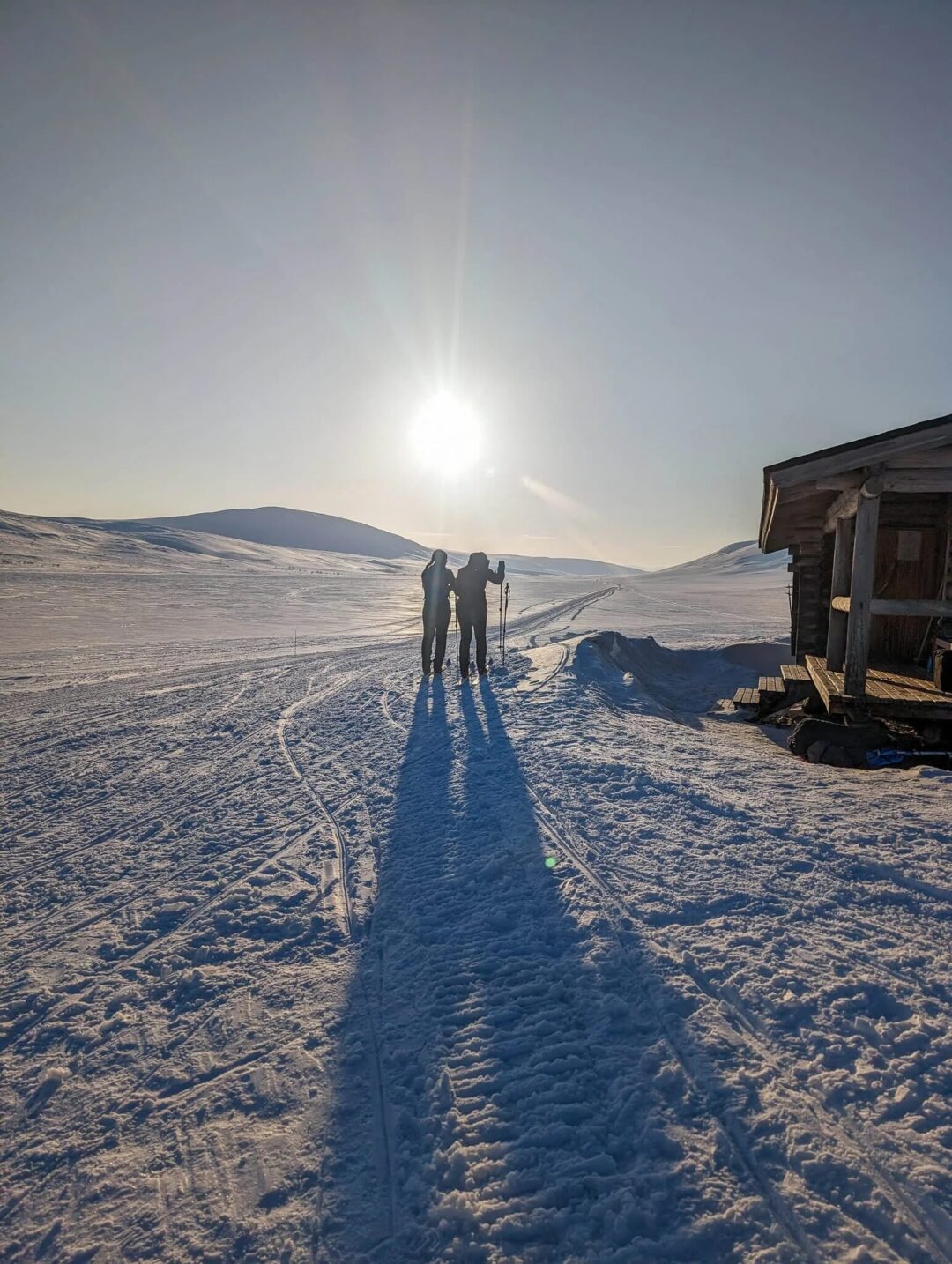 Two skiers walking a snow trail past an Arctic hut, heading across the Arctic tundra in clear winter light