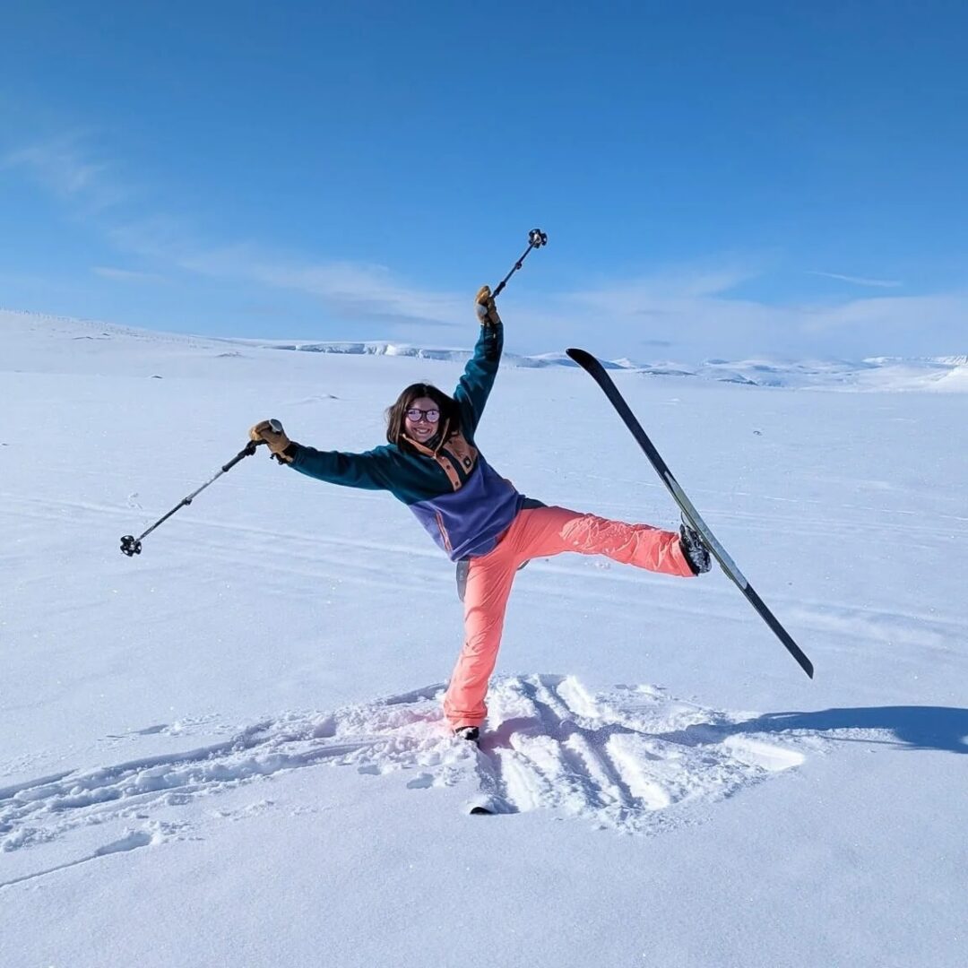 Skier celebrates mid-air on an Arctic ski trek across snowy white plains with poles under clear blue sky