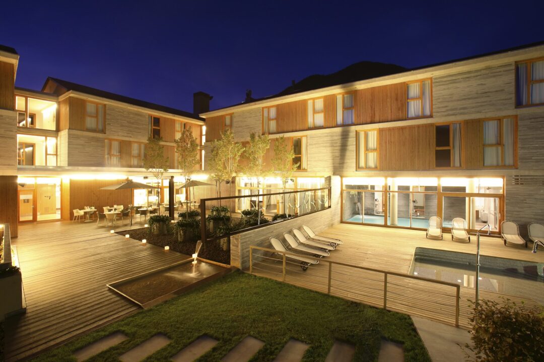 Lit wooden lodge with balconies and terrace courtyard at night, with loungers and patio lights
