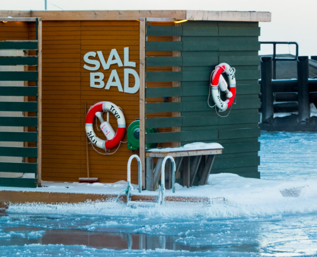 Lifebuoys and “SIAL BAD” sign outside a winter swimming pool by snowy water and an inflatable safety device