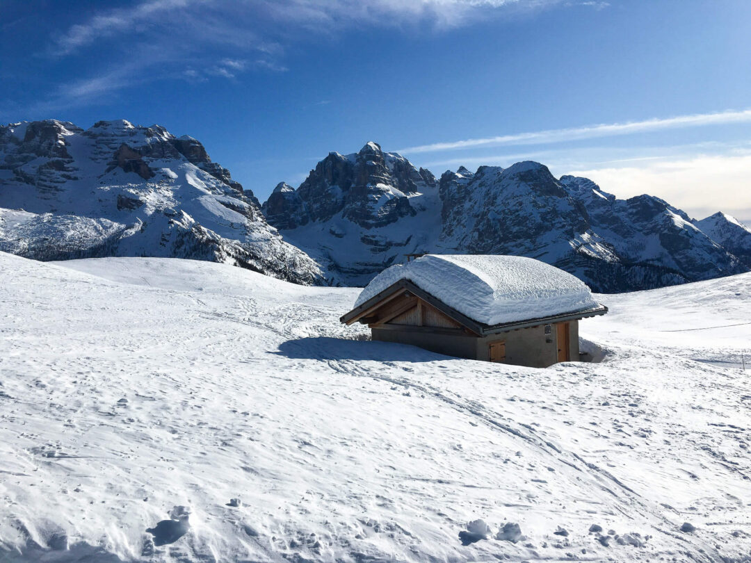 Snow-covered Alpine hut in the Dolomites mountains under a blue sky, with ski slopes and peaks beyond