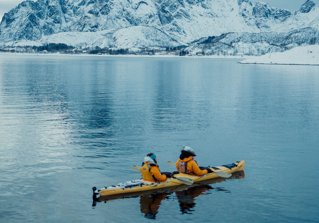 Two kayakers paddle on a Lofoten fjord, with snowy mountains and glaciers in the background.