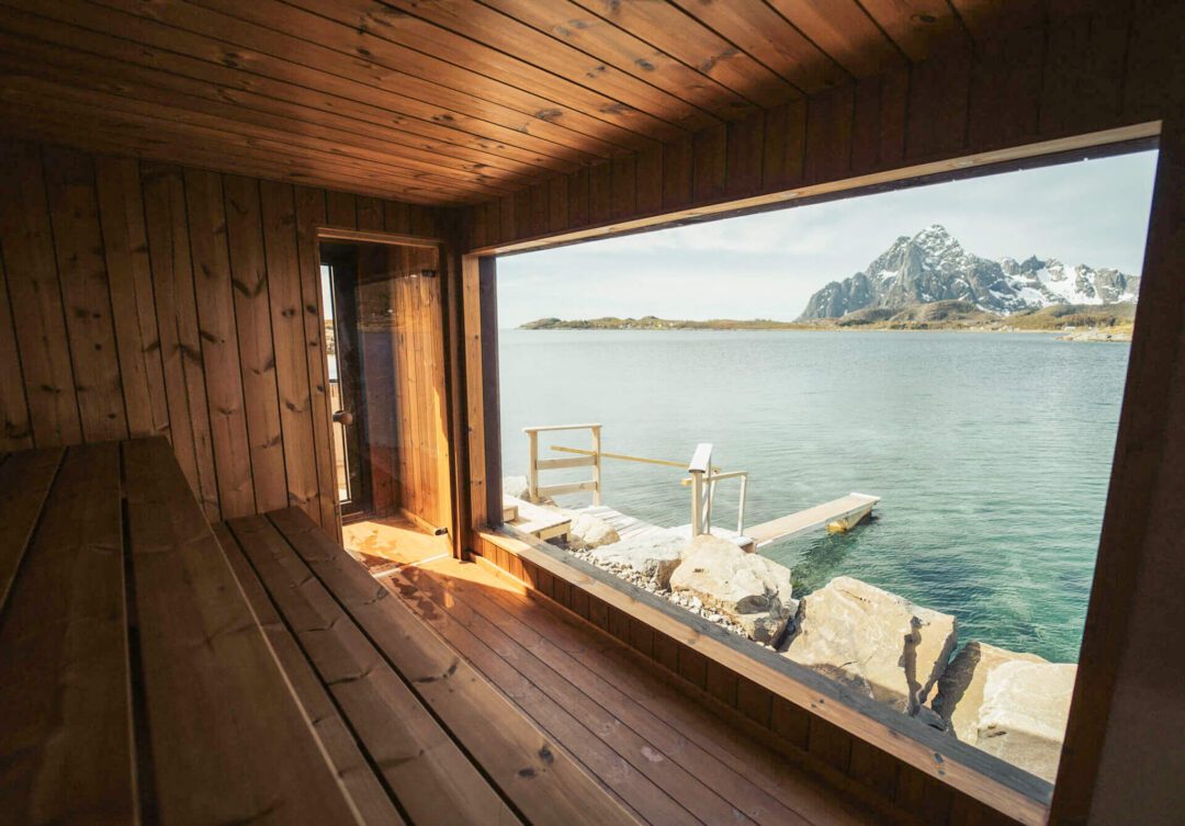 Coastal sauna window with sea view and Lofoten mountains in the distance, wooden interior and rocky waterfront