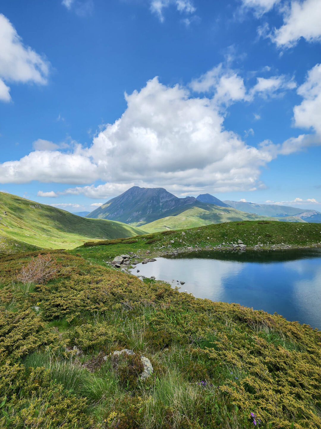 Scenic hiking landscape with mountain lake, green grassy hills and peaks under a blue cloudy sky.