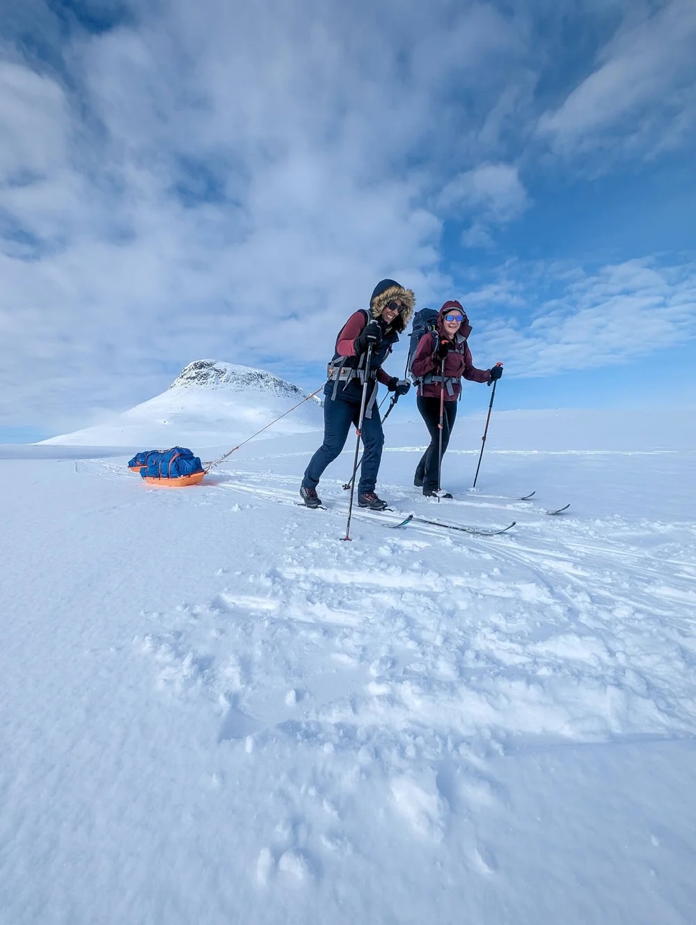 Two skiers with backpacks and poles hiking across snow on a high Arctic ridge under a blue sky