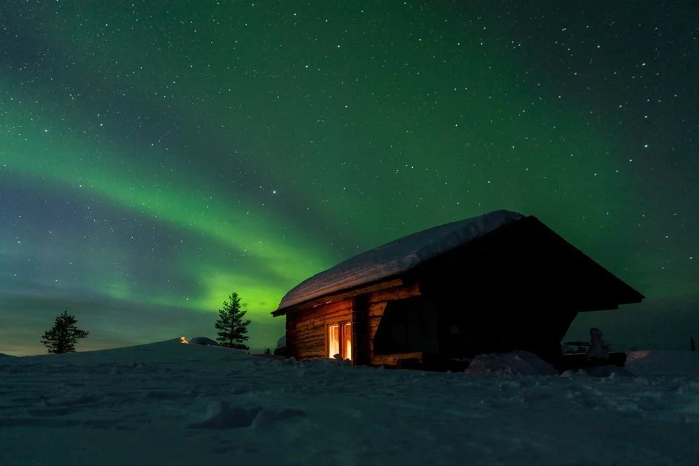 Aurora borealis over a remote Arctic ski hut at night with warm light glowing in snowy landscape