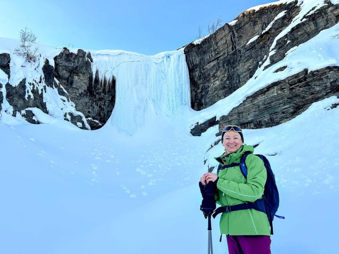 Skier smiling in front of frozen waterfall in snowy Arctic mountains during a ski expedition