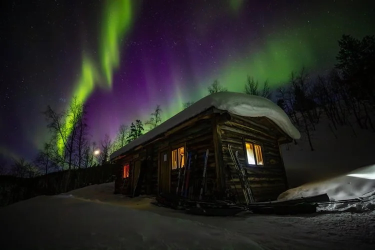Aurora borealis over a snow-covered log hut on an Arctic ski expedition route at night