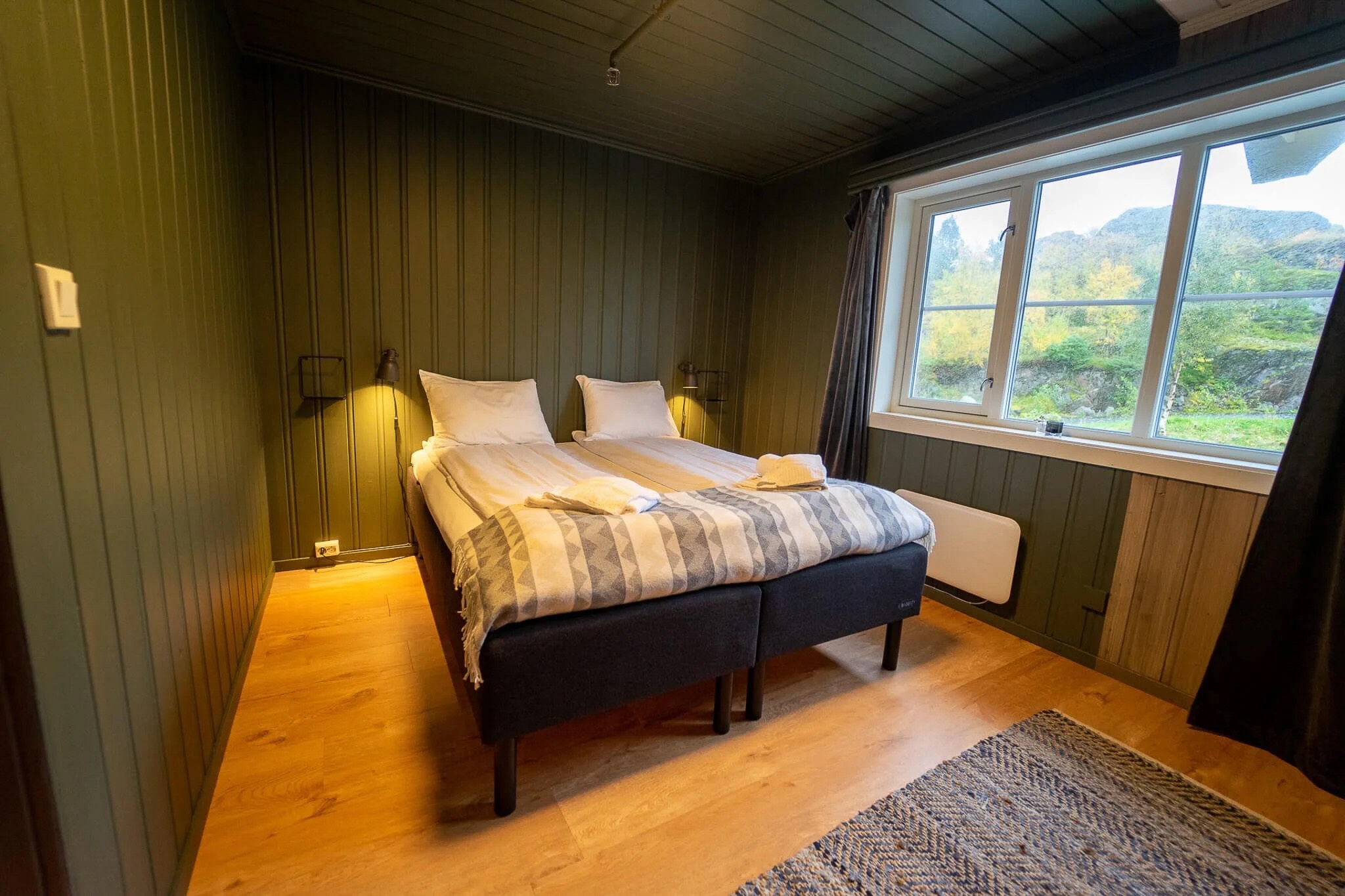 Cozy cabin bedroom with double bed and large window overlooking green mountains in Lofoten