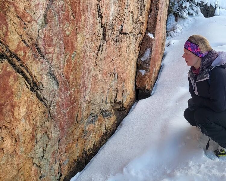 Woman in winter gear kneels beside a rocky wall on a snowshoe trail in Finland.