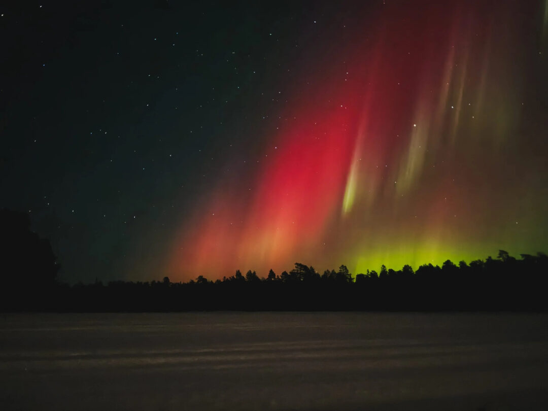 Northern lights (aurora borealis) over a snowy Finnish landscape at night, with green and red streaks across the sky