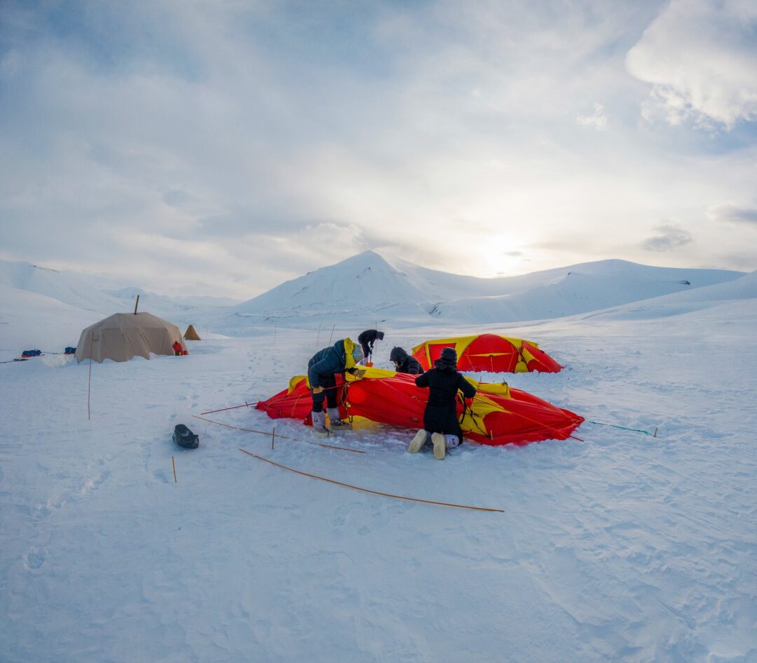 Women’s Arctic wild camping campsite in Svalbard, with tents and supplies in snowy terrain at dusk.