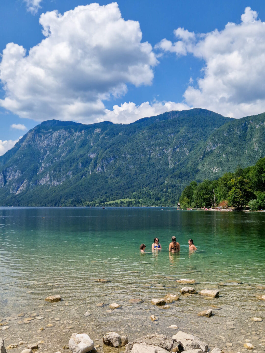 Women wading in a calm lake with forested mountains under blue sky in Slovenia