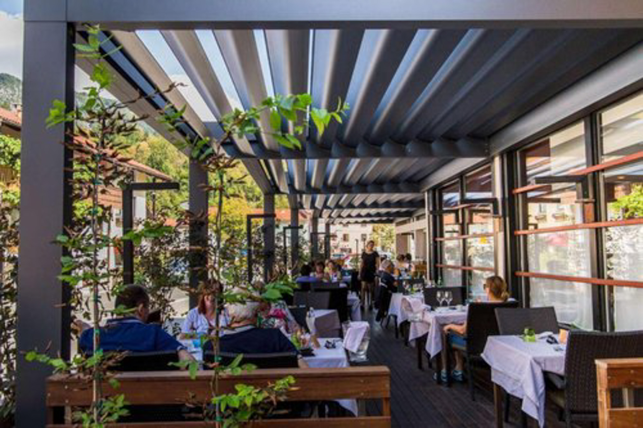 Outdoor dining terrace with tables and potted plants under a metal canopy.