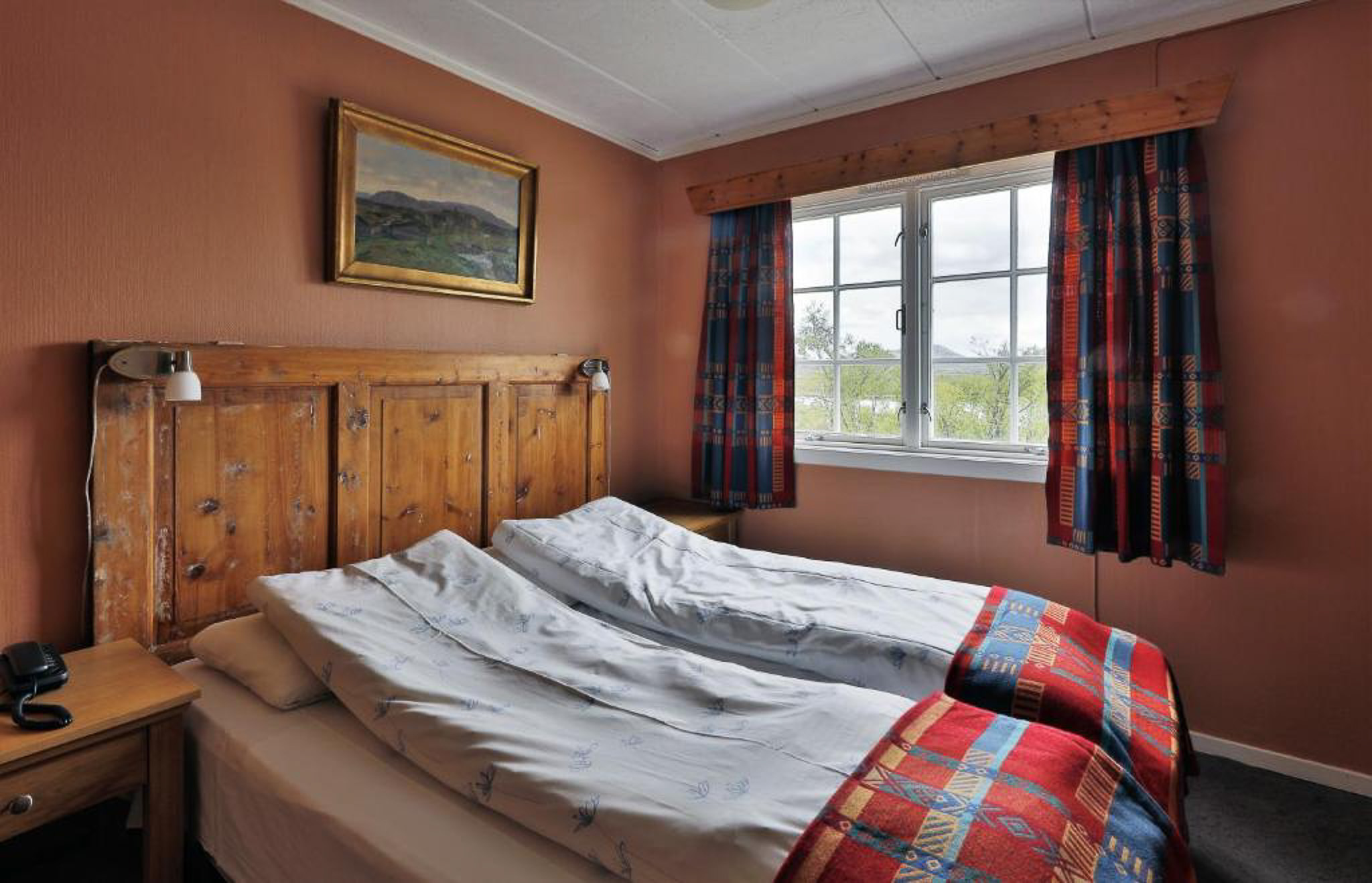 Unmade double bedroom with wooden headboard, window with curtains, and landscape painting on the wall