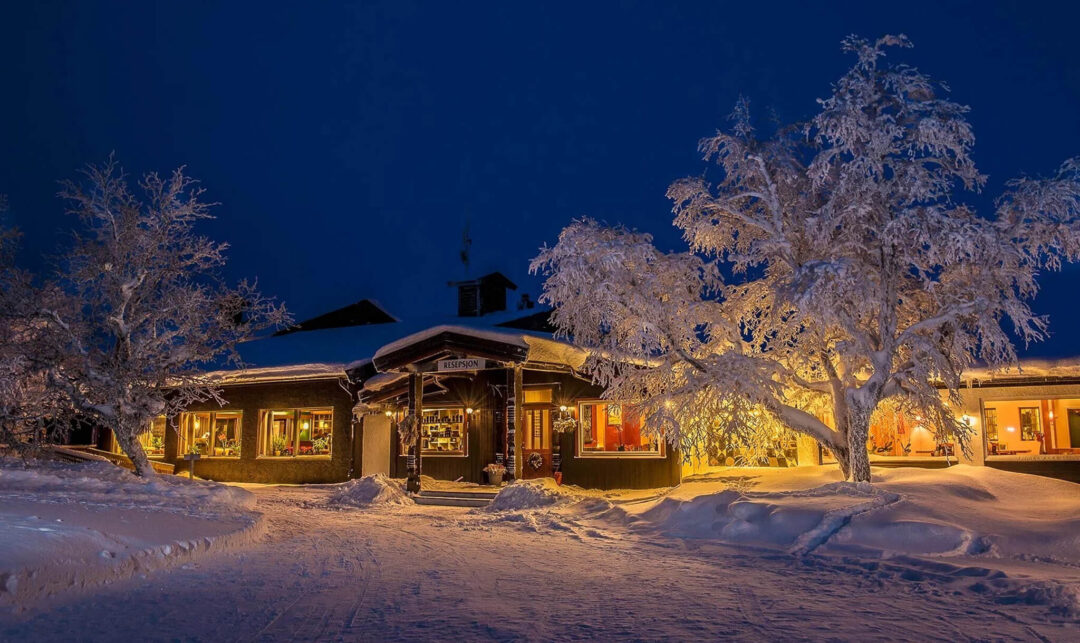 Snow-covered wooden cabin at night with warm lights and pine trees, winter landscape