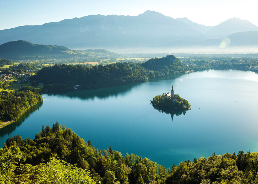 Aerial view of a Slovenian lake with forested island and church, surrounded by mountains and mist