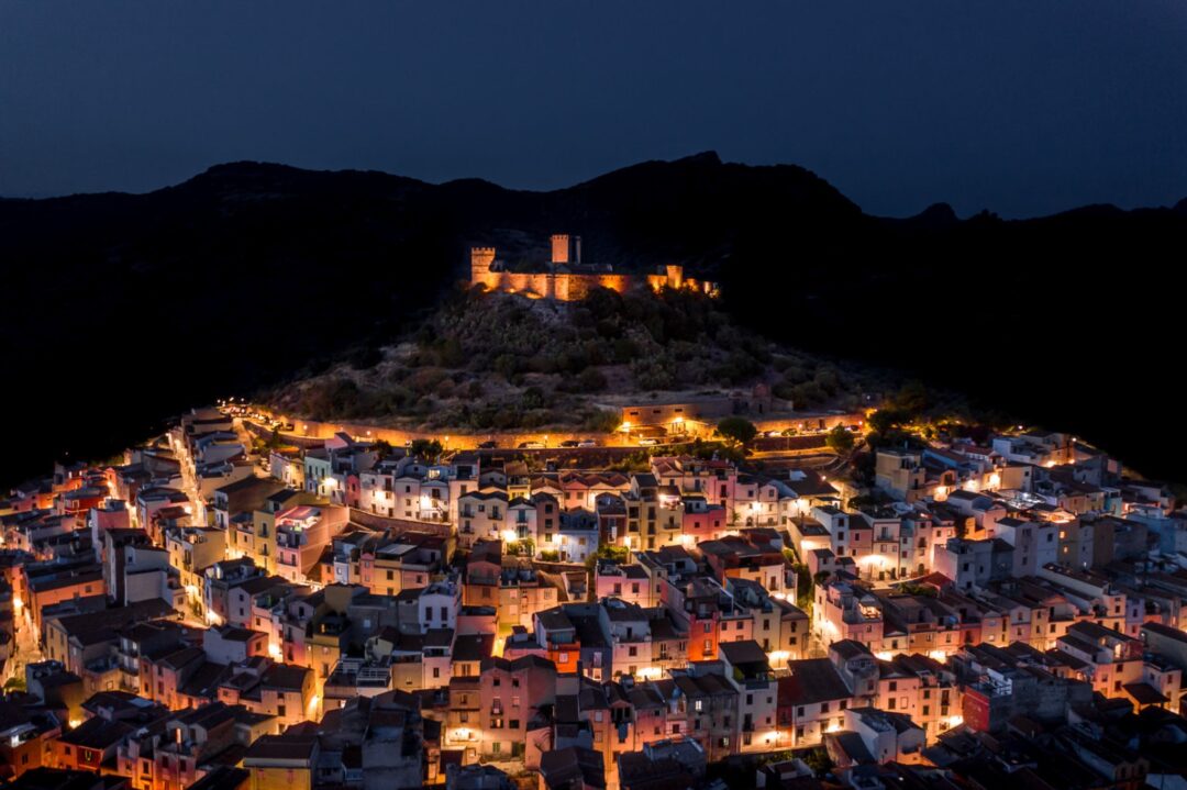 Night view of a coastal hill town with warm-lit buildings along a dark mountain ridge