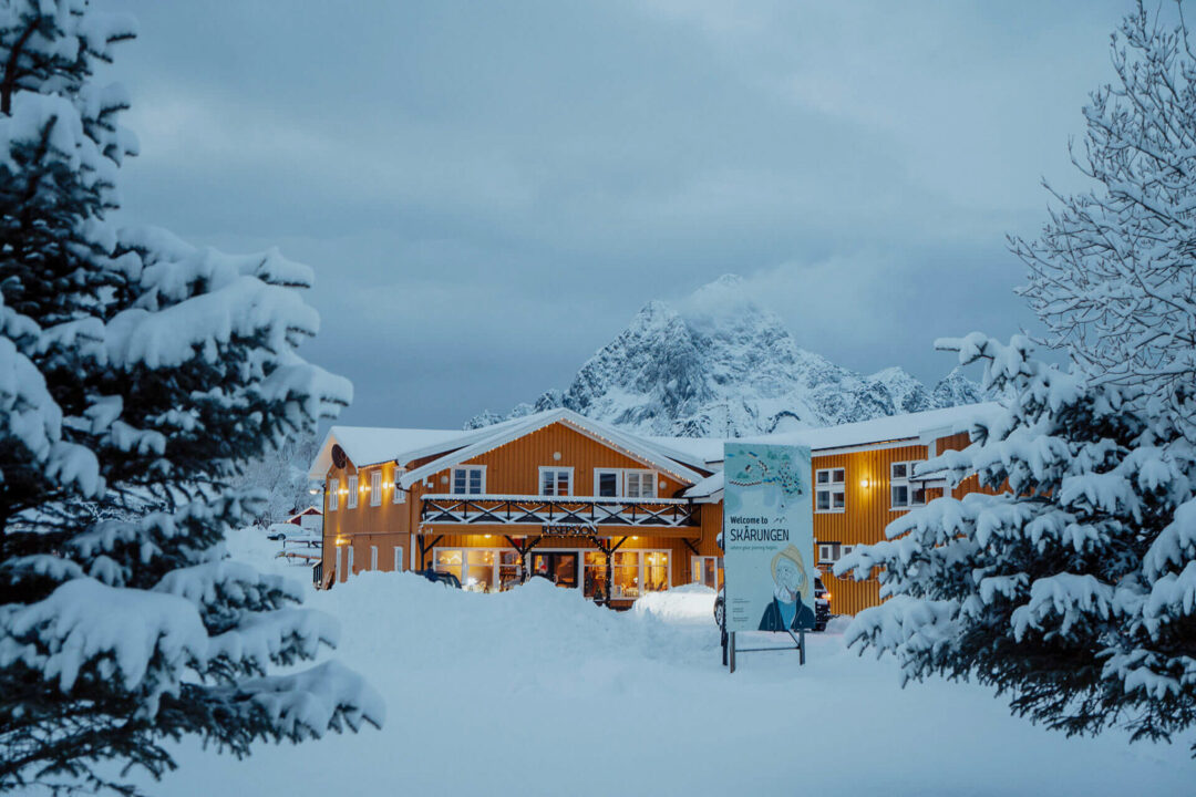 Snowy mountain lodge with warm lights and a sign in the foreground, surrounded by pine trees and deep snow