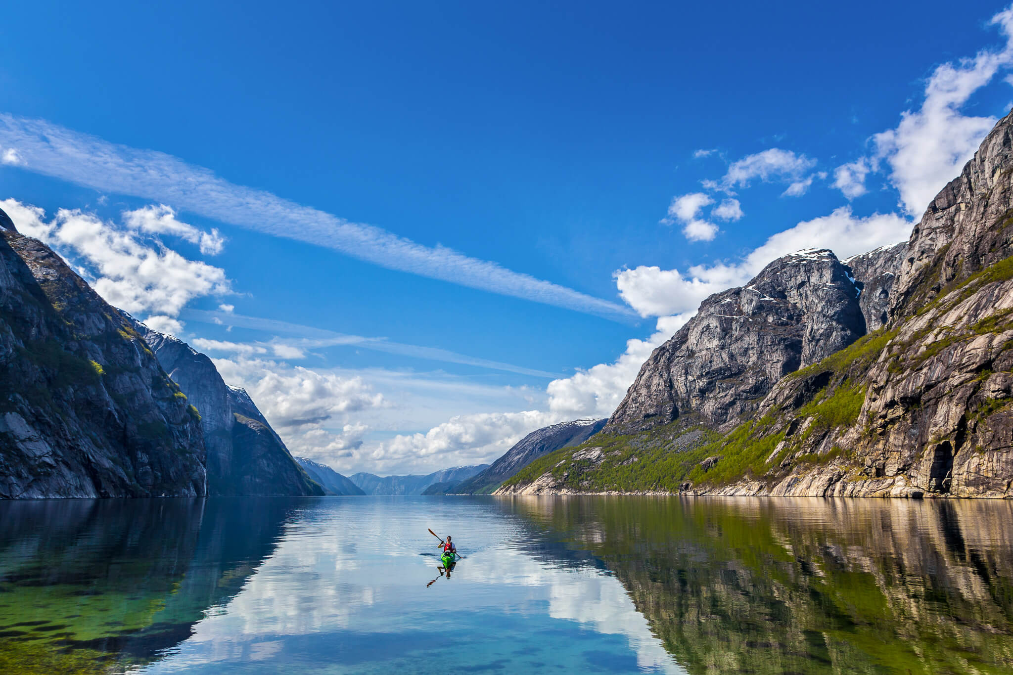 Kayaker paddles through a calm mountain fjord with steep cliffs and blue sky reflections