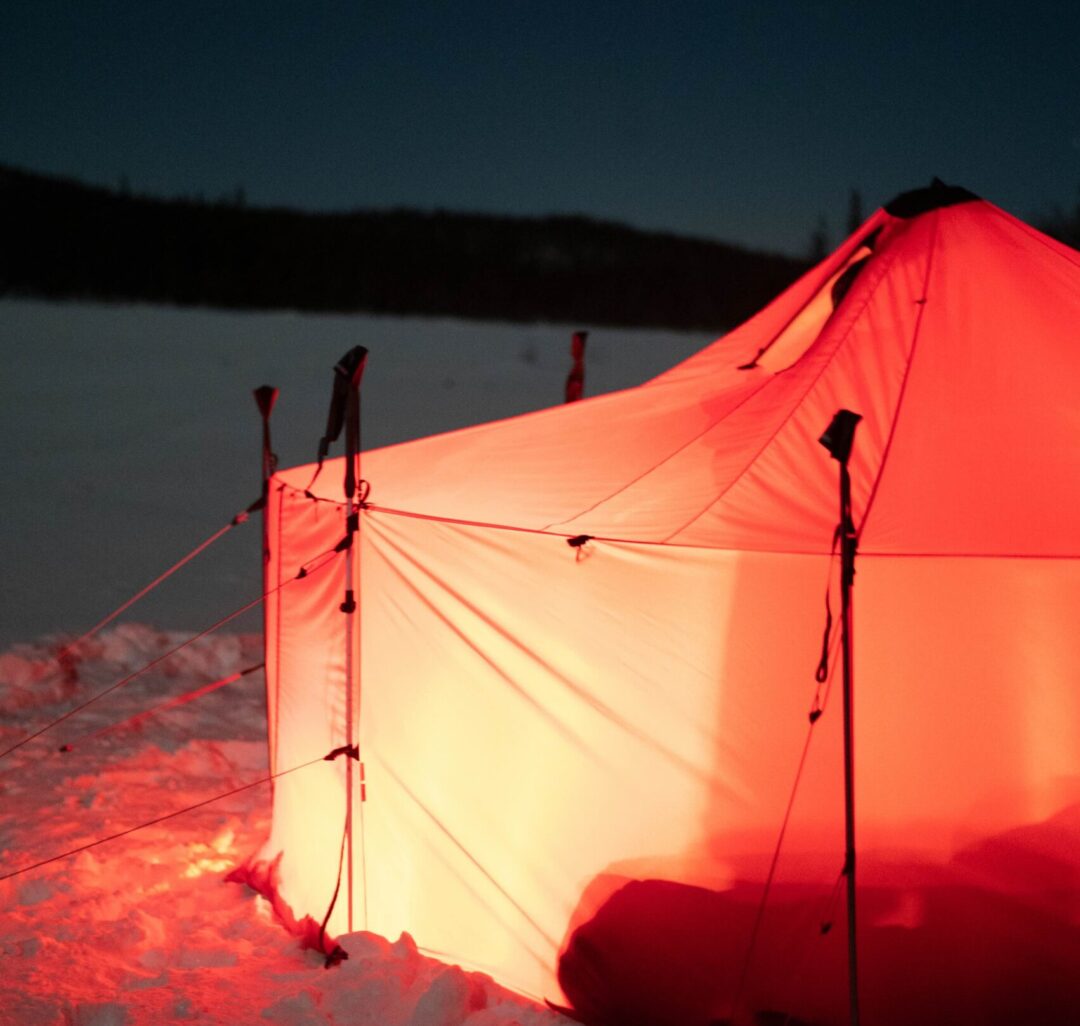 Red winter camping tent pitched on snowy ground at dusk, with guy ropes and tent poles.