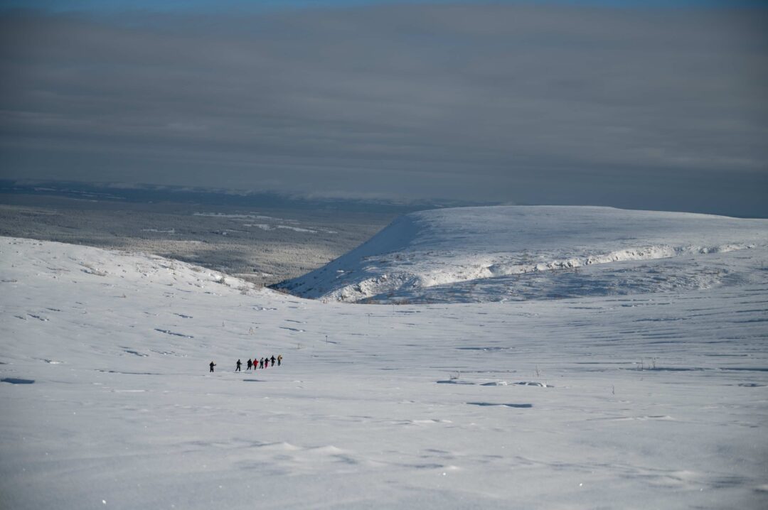 Skiers traverse snowy backcountry slopes across a winter mountain valley under a cloudy sky