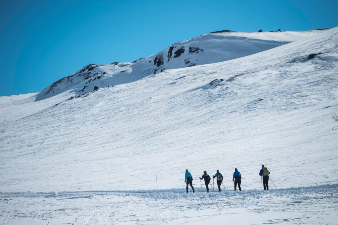 Women cross-country skiing on snowy slopes with mountain peaks in Norway under a clear blue sky