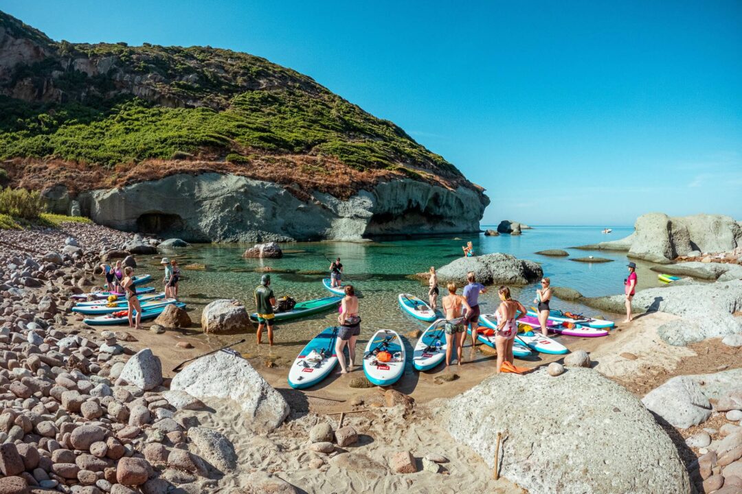 Women on paddleboards at a SUP beach with kayaks near cliffs in Sardinia, with others in calm blue water