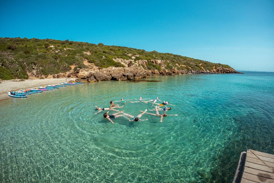 Women doing SUP on calm turquoise water off the Sardinia coast, with boats at the shoreline