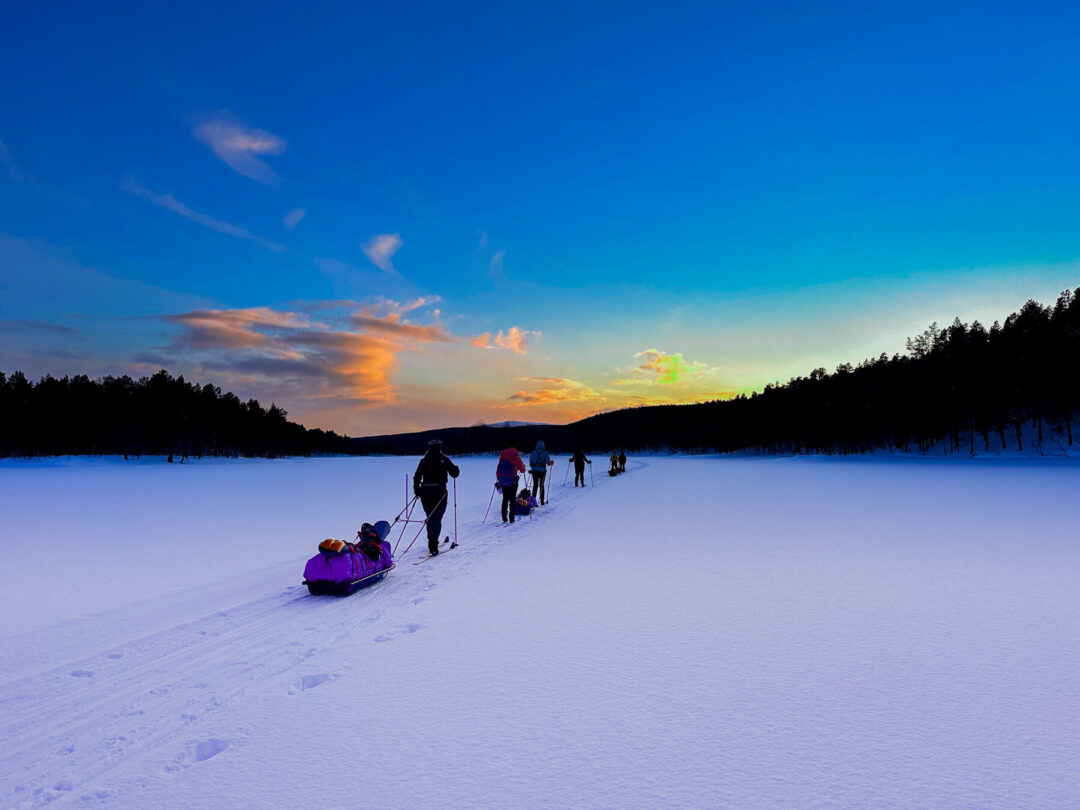 Cross-country skiers travel across a snowy lake and forest at dusk on a winter expedition, pulling sleds.