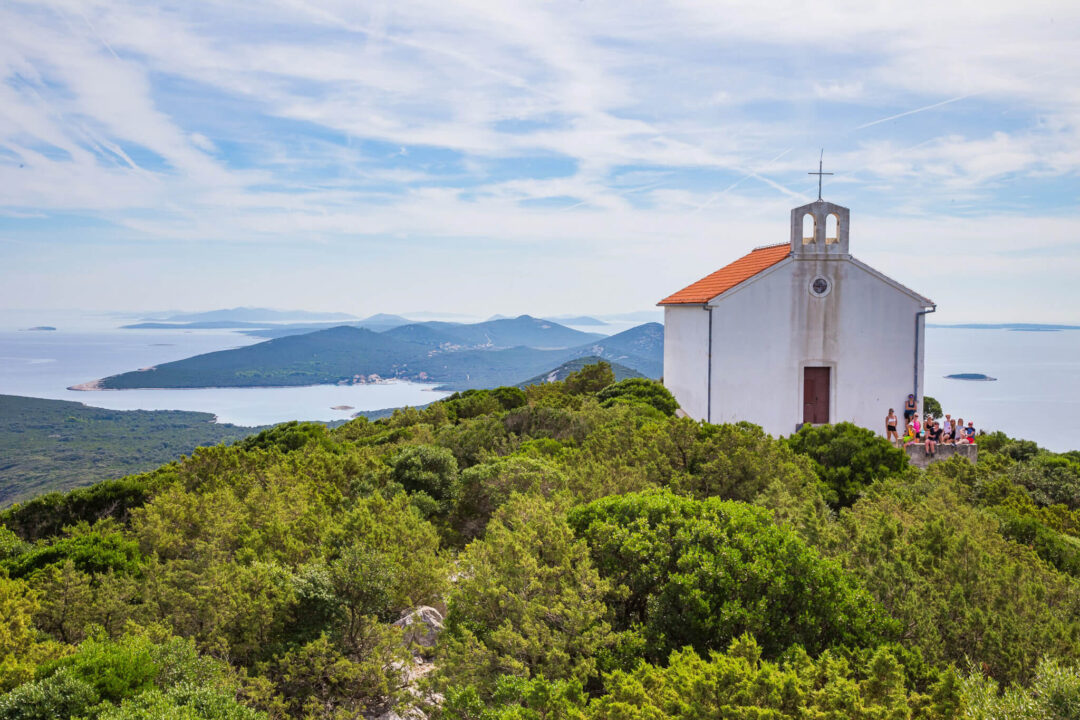 Women walk beside a small white chapel overlooking the sea from a green island