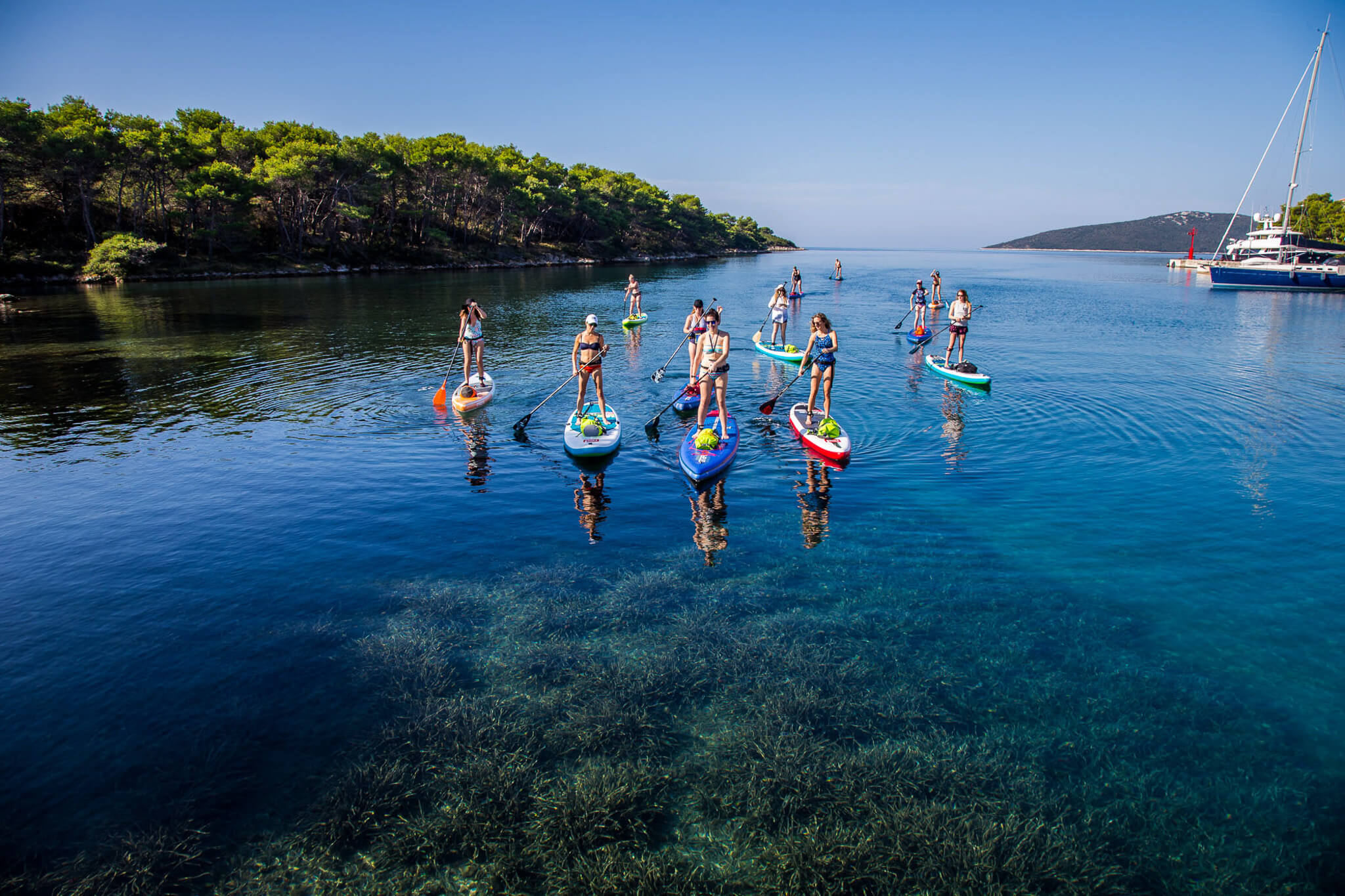 Women stand-up paddleboarding in a coastal bay, with a sailboat offshore.