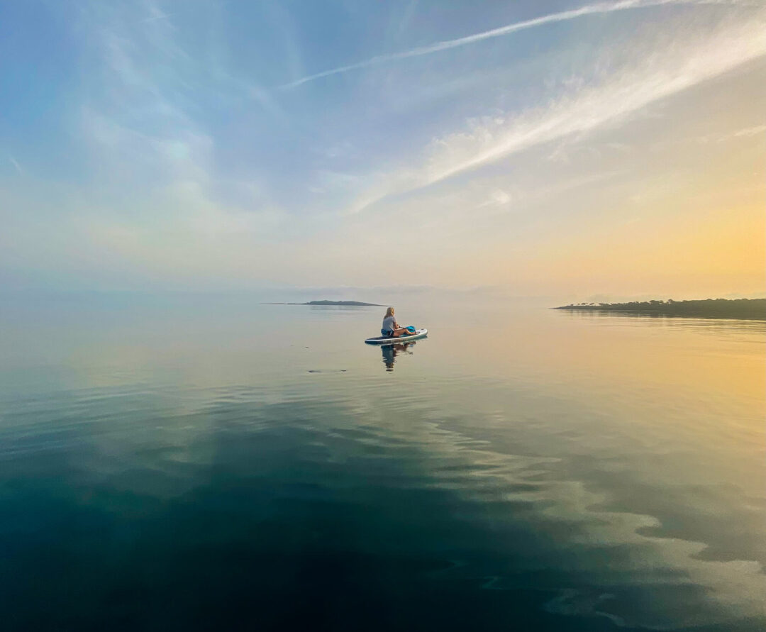 Woman paddleboarding on calm water at sunset, seen from a low viewpoint