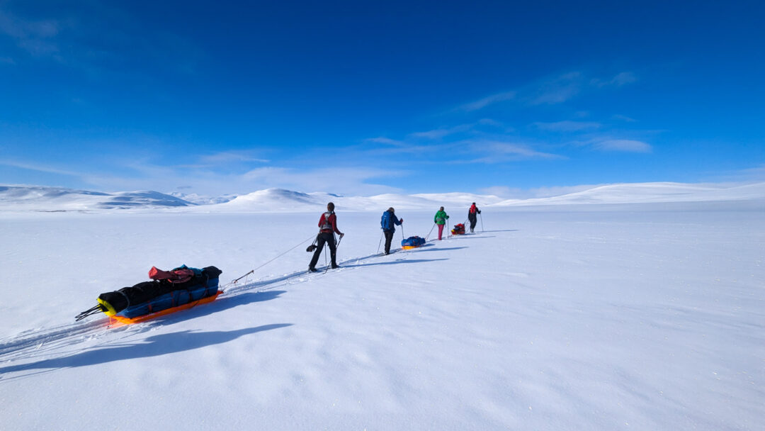 Cross-country ski expedition across snowy mountains with sleds, distant skiers on a ridge under blue sky