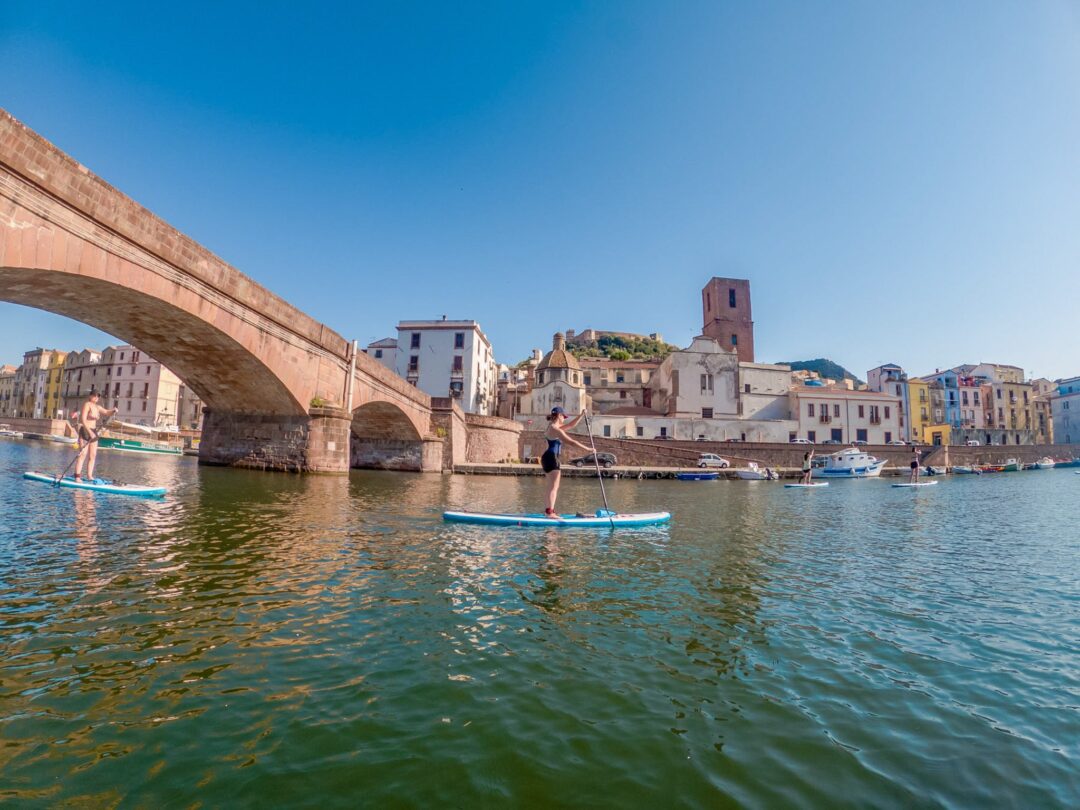 Women paddleboarding on calm water in a harbour, with old buildings and arches in Sardinia