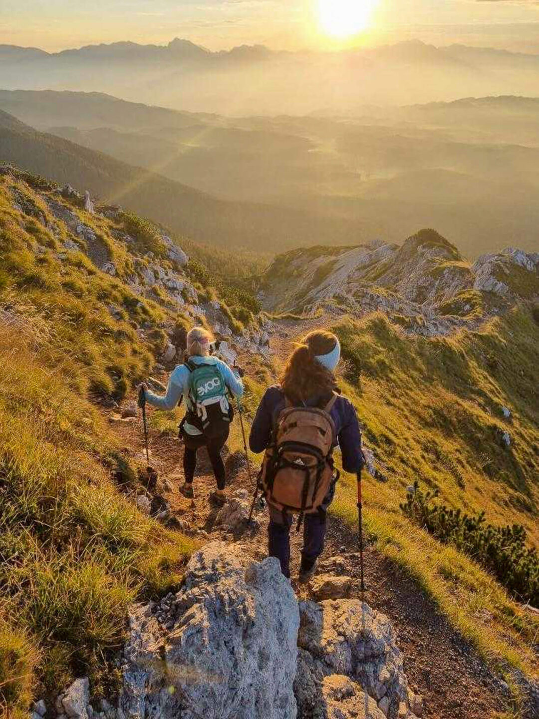 Women hiking along a mountain trail at sunrise in Slovenia, with huts-to-huts adventure scenery in the valley.