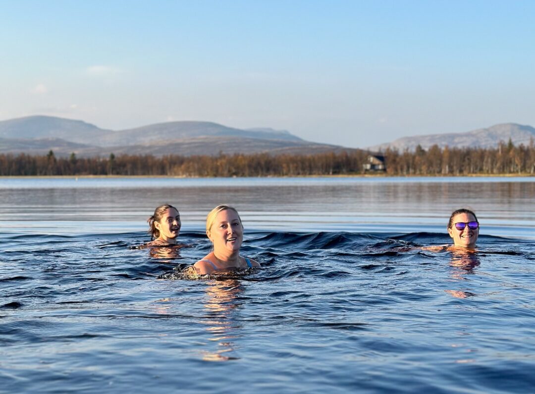 Three women swimming in a lake with trees and rolling hills in the background at sunrise