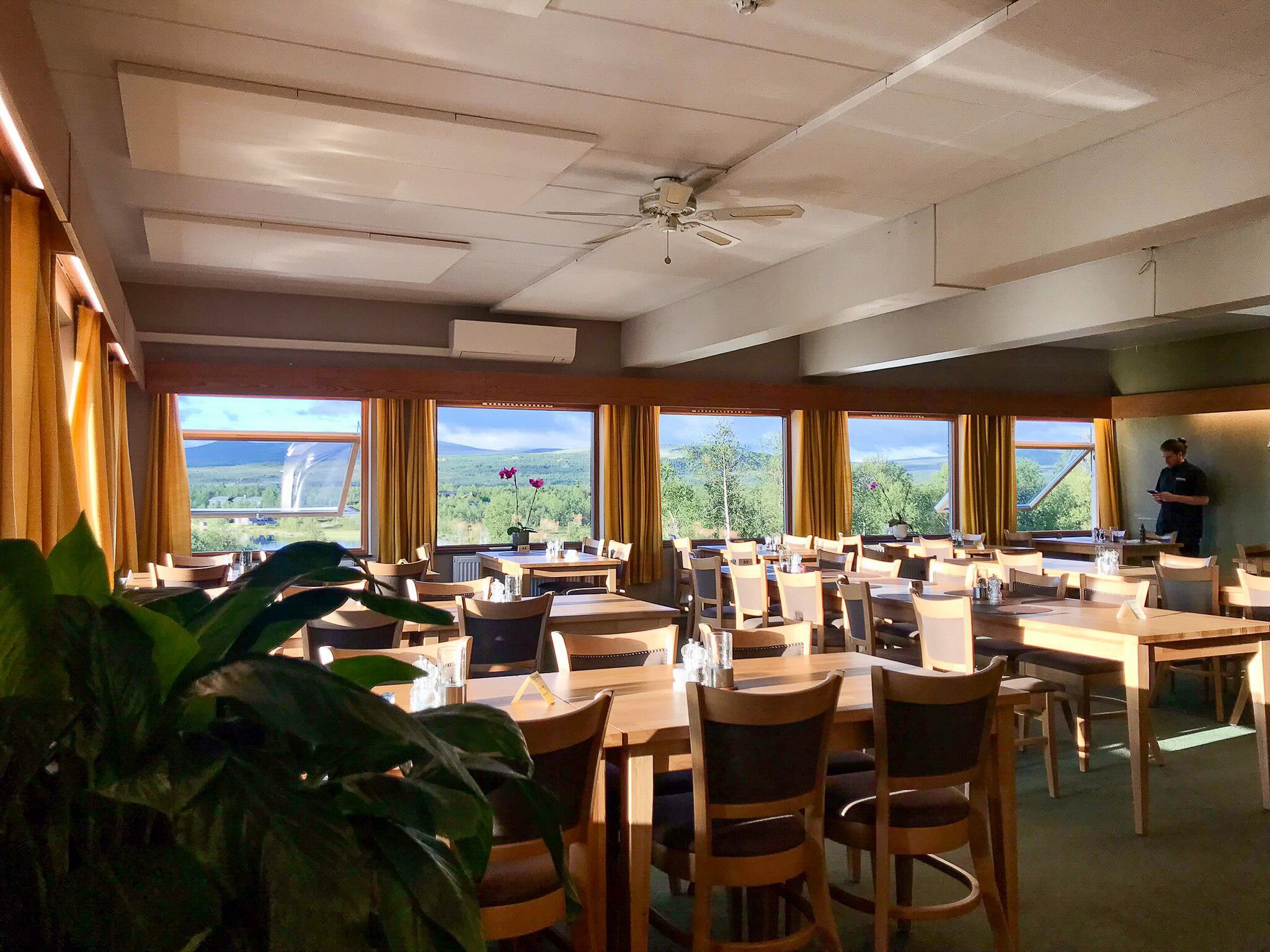 Bright dining room with tables and chairs, large windows and scenic countryside views, with a man standing by a wall