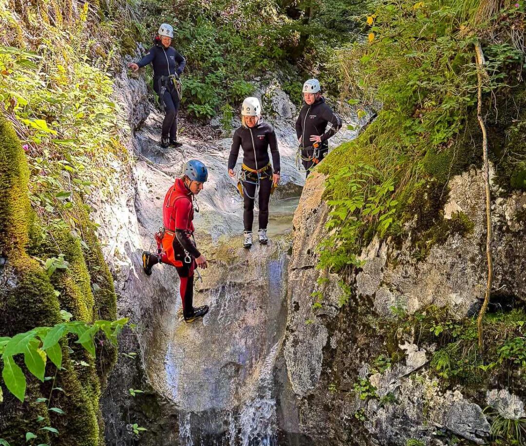 Women in wetsuits and helmets walk through whitewater in a rocky river gorge canyon