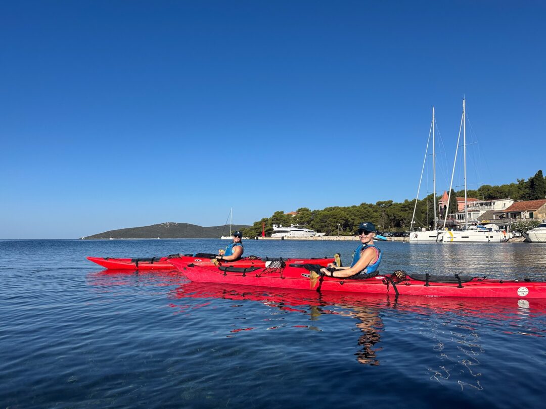 Women kayaking on calm sea near a marina, with sailboats and coastal houses in the background.